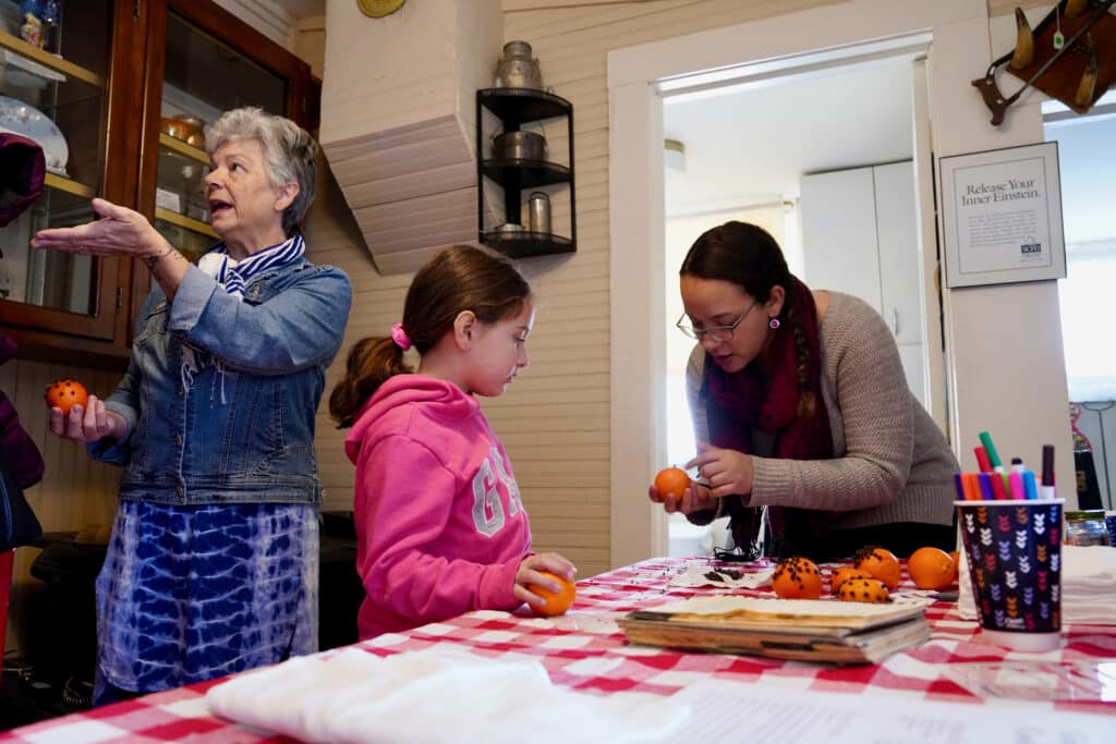 A young girl makes a pomander with the help of a museum staff. Two adults chat in the background.