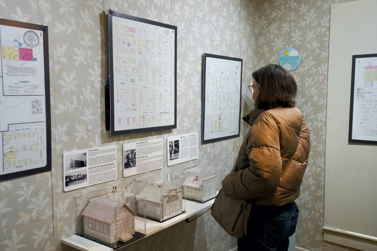Photo of woman in brown jacket looking at historic maps hanging on a wall. On a shelf below the maps are models of buildings made out of paper.